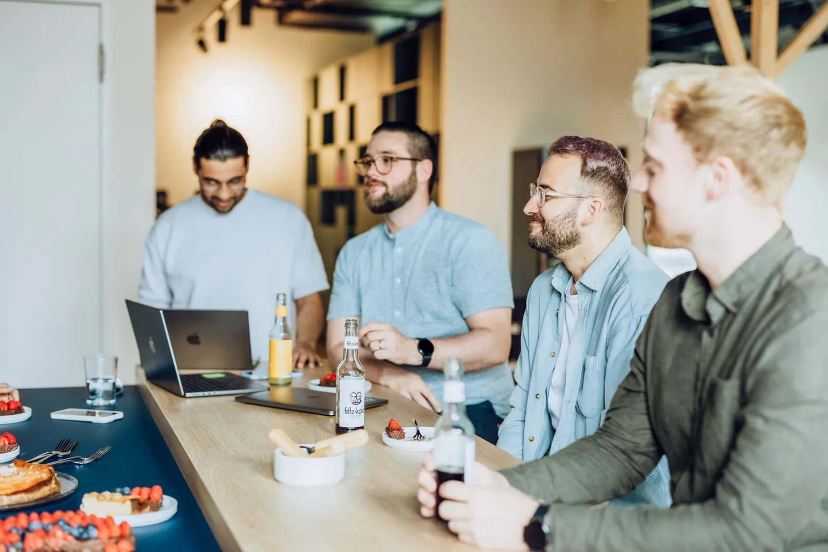 Four men sitting at a table with laptops and drinks, enjoying a casual meeting with desserts.
