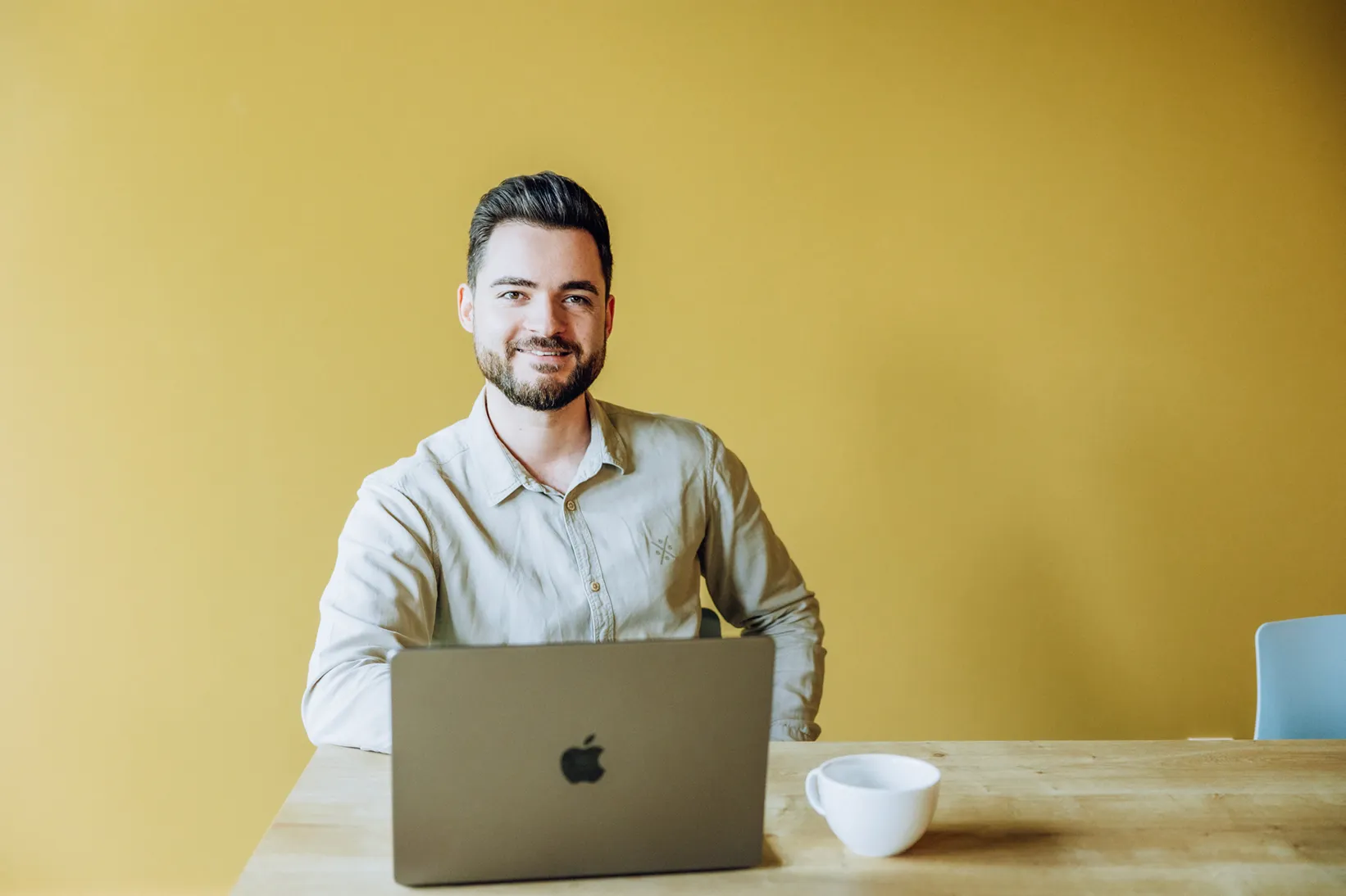 Man sitting at a table with a laptop and a coffee cup, smiling against a yellow background.