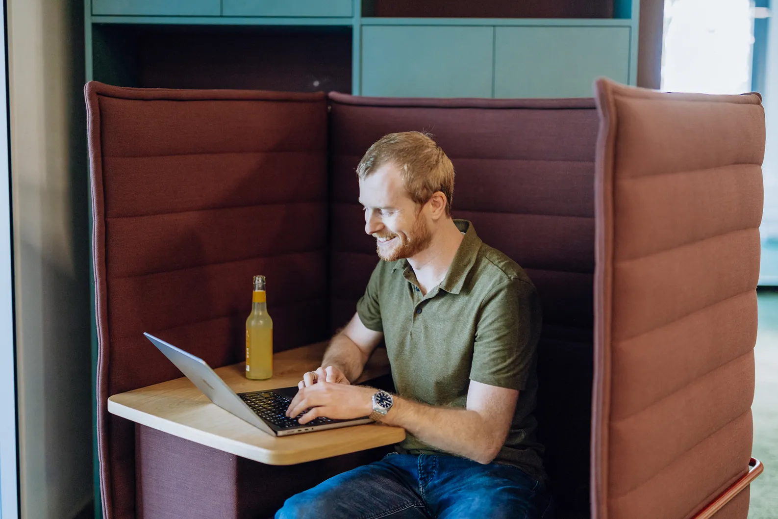 A man sitting in a cozy booth working on a laptop with a bottle of drink on the table.