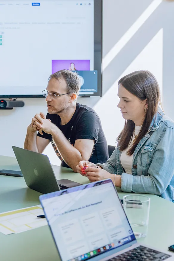 Two people sitting at a table with laptops, engaged in a discussion, with a screen displaying content in the background.