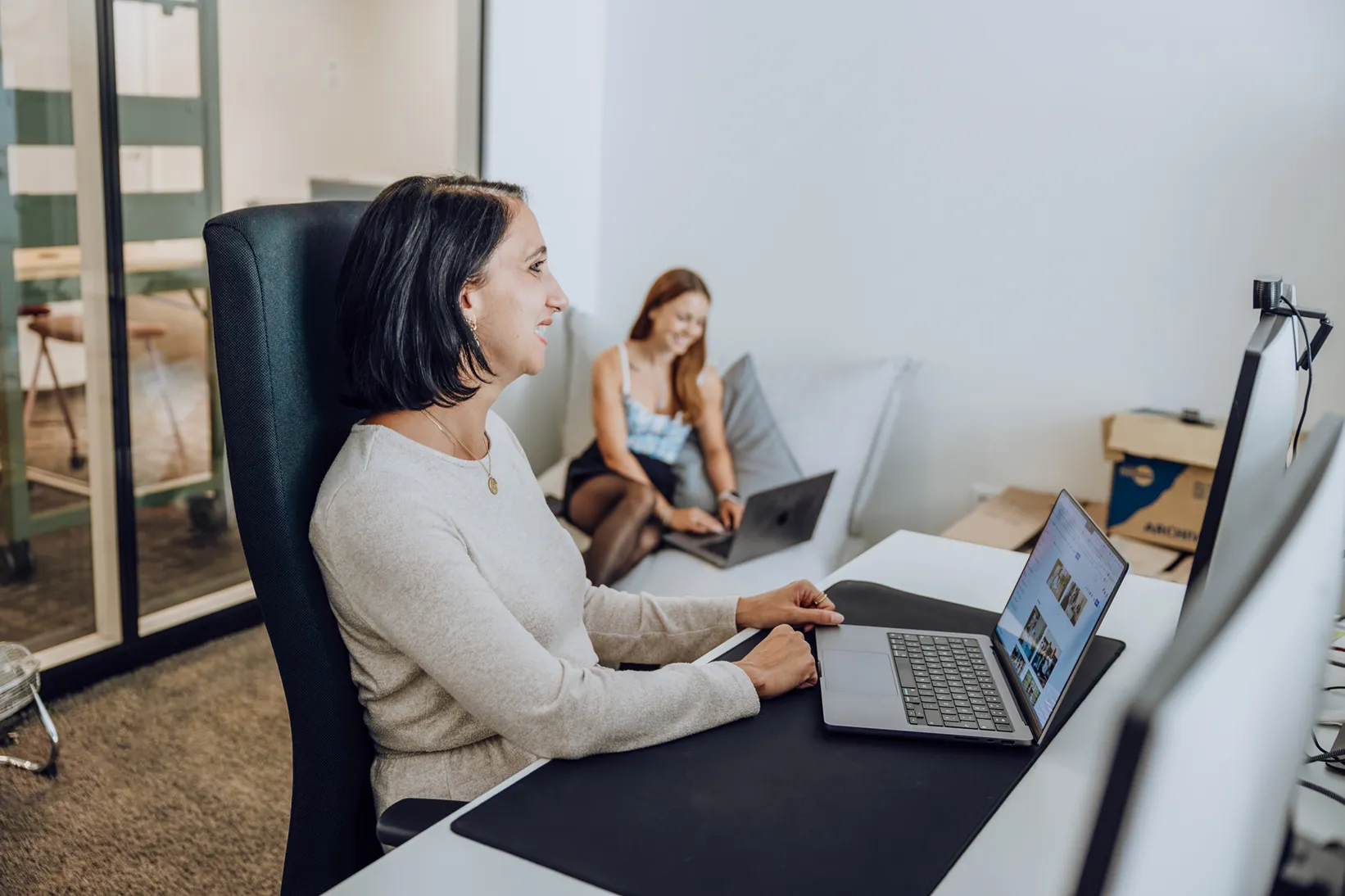 Two women working on laptops in a modern office setting, one sitting at a desk and the other on a couch.